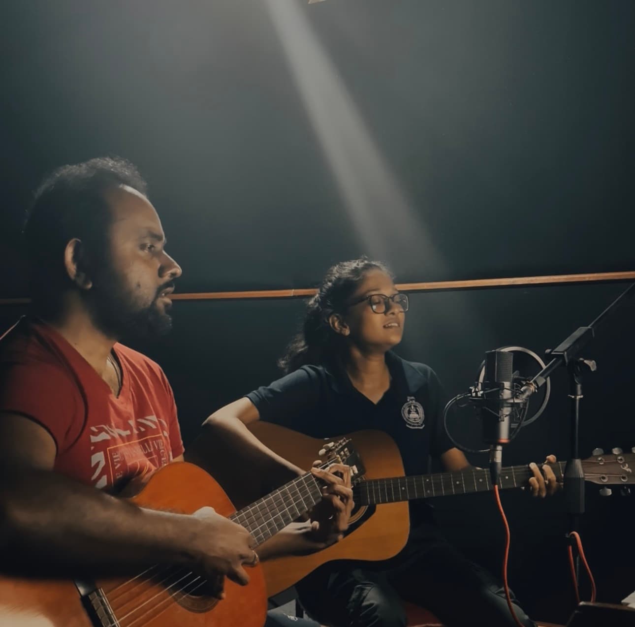 Two performers playing acoustic guitars and singing into a studio microphone under a focused beam of light.